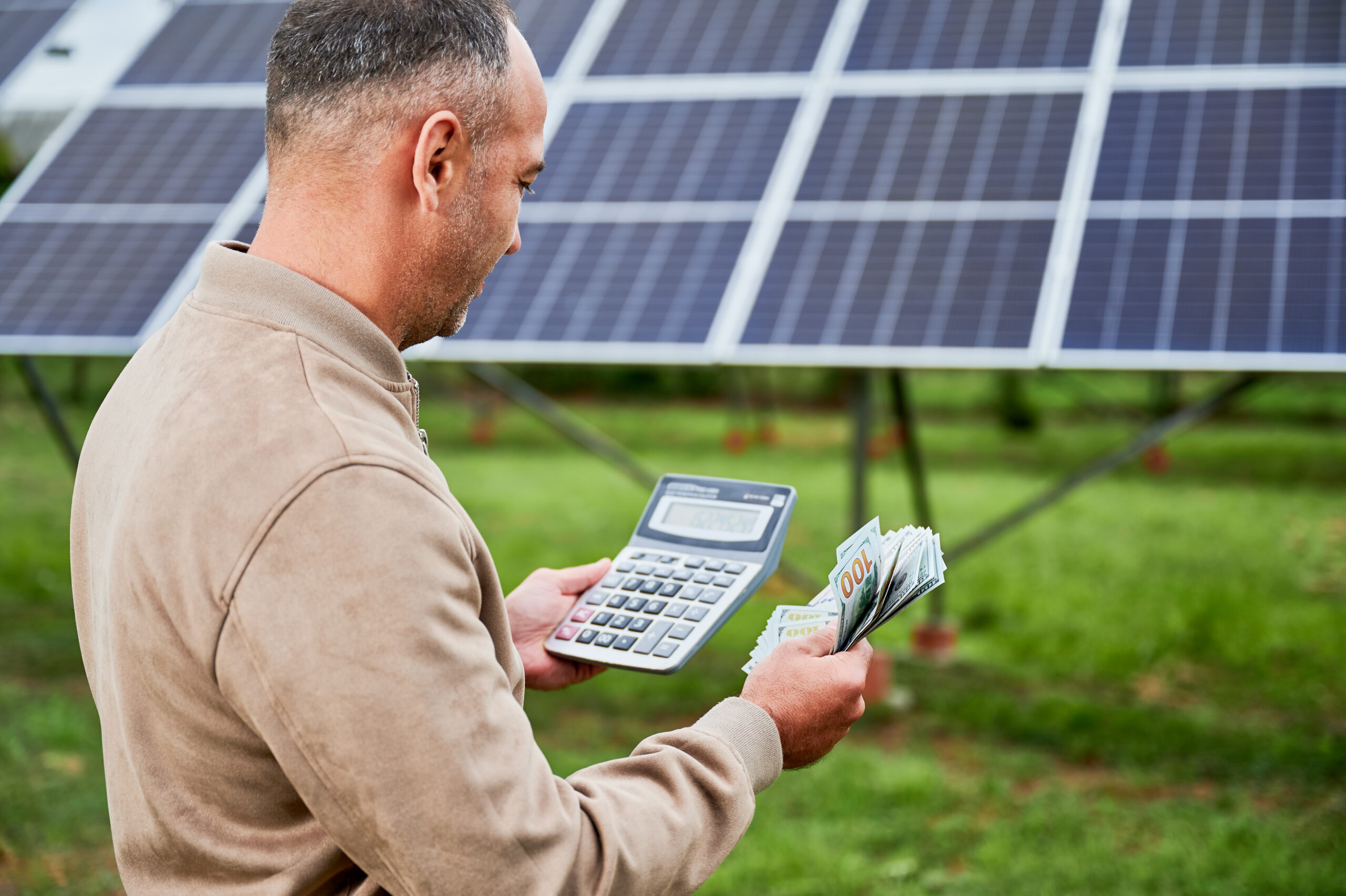 Confident investor counting profit he got from investing in green energy. Glad man holding huge sum received by successfull investment. Back view of man with calculator and money next to PV battery.